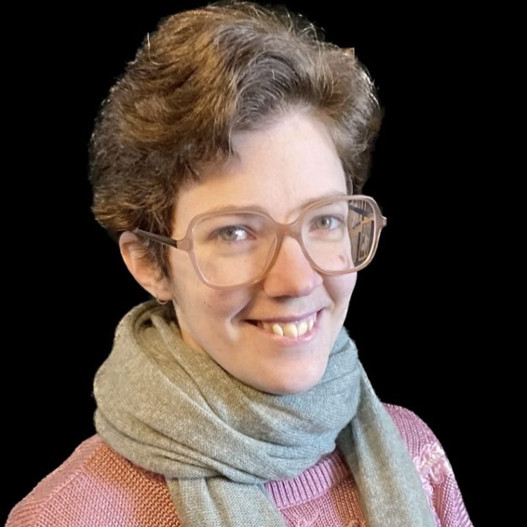 Junge Frau mit lockigem, kurzem Haar, Brille und einem grauen Schal, lächelt freundlich.