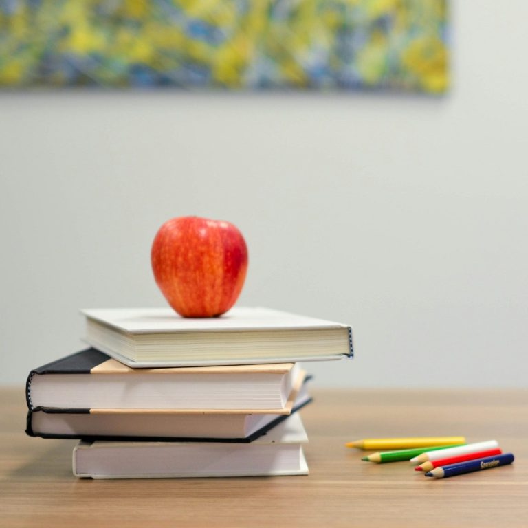 Rote Apfel auf gestapelten Büchern neben Buntstiften auf einem Tisch.