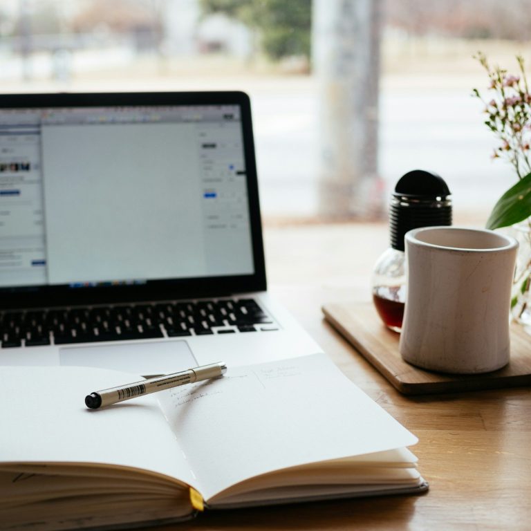 Laptop mit offenem Notizbuch, Kaffeetasse und Blumen auf einem Tisch.