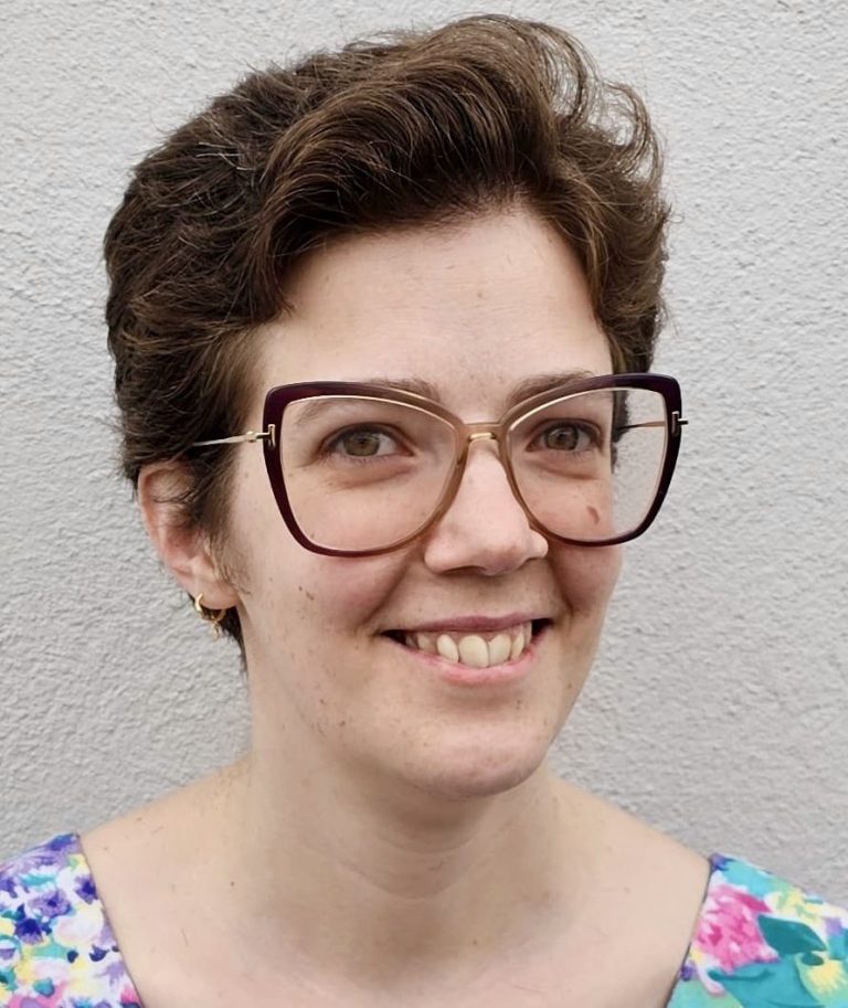 Eine Person mit kurzer brauner Frisur, Brille und grauem Schal, lächelt freundlich.