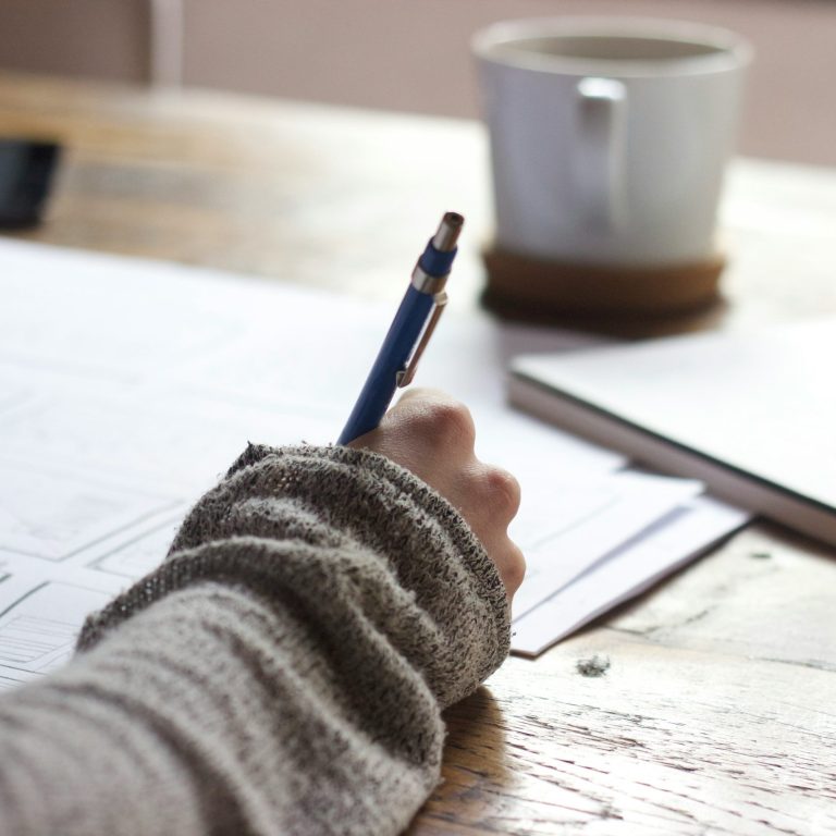 Eine Hand mit einem Stift, die auf ein Blatt Papier schreibt, neben einer Tasse.