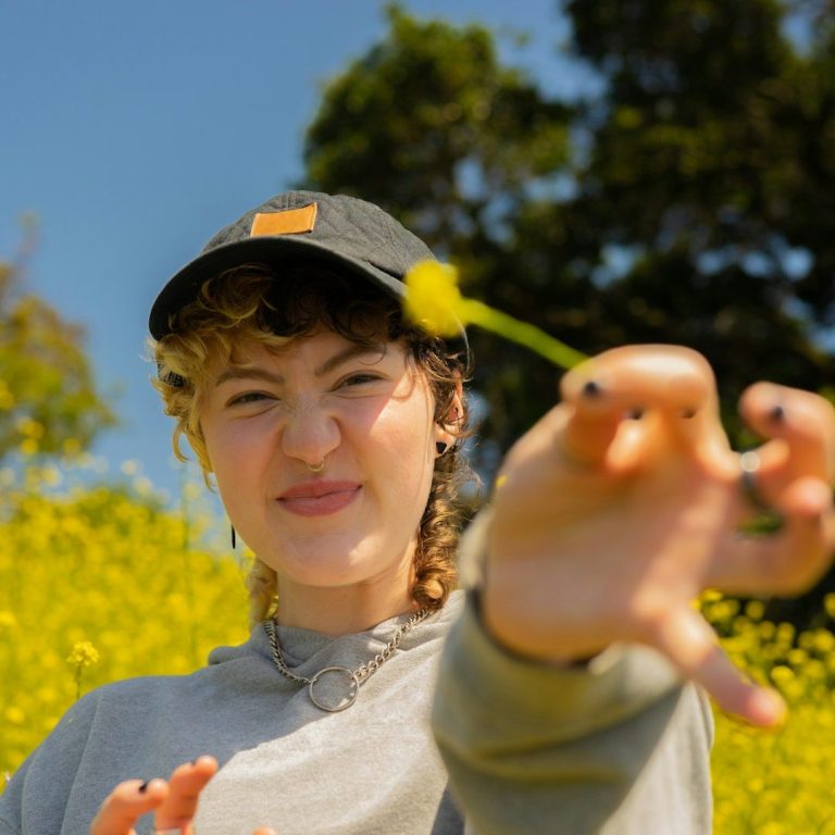 Junge Frau in einem Feld voller Blumen, die eine Blume in der Hand hält und lächelt.