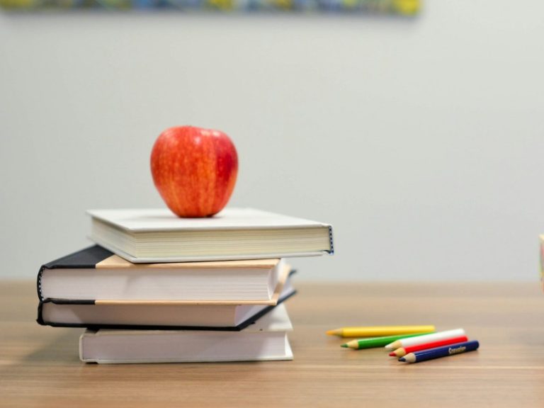 Ein roter Apfel auf einem Stapel Bücher neben bunten Stiften.